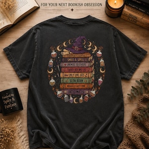 May include: A charcoal grey t-shirt featuring a graphic on the back of a stack of books topped with a purple witch hat. The books are labelled with text including Smut & Spells, Enemies to Lovers, and Fated Mates, surrounded by potion bottles.