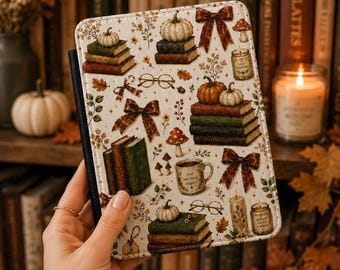 May include: A rectangular e-reader case featuring a cream-colored background with a repeating pattern of stacked books, pumpkins, brown bows, eyeglasses, mushrooms, candles, and a coffee mug with the text "Just one more chapter".