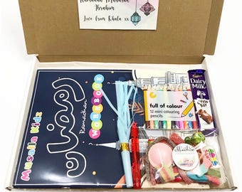 May include: An open cardboard box filled with children's Ramadan gifts. Includes a navy blue workbook, colorful pencils, candy, and a chocolate bar. The box has a decorative card that says "Ramadan Mubarak Ibrahim".