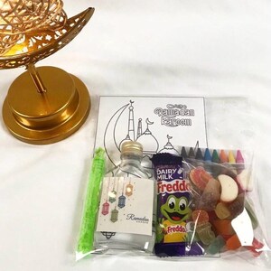 May include: A clear cellophane bag contains a small bottle, a chocolate bar, a coloring book, crayons, and gummy candies. The coloring book has a mosque illustration and the words "Ramadan Kareem". A gold-colored decorative lamp is in the background.