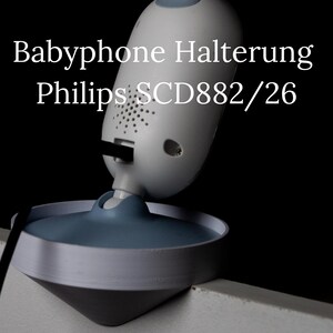 Könnte beinhalten: Ein weiß-grauer Babyphone-Monitor mit dem Text "Babyphone Halterung Philips SCD882/26". Der Monitor hat eine abgerundete Oberseite mit einem Lautsprecher und eine Basis mit blauem Rand. Ein schwarzes Kabel geht vom Monitor ab.