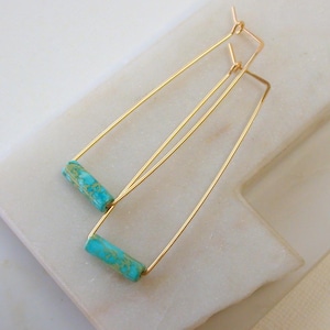 May include: A pair of gold-colored, geometric earrings. Each earring features a turquoise-colored, cylindrical stone at the base of a long, triangular frame. The earrings are displayed on a white surface.