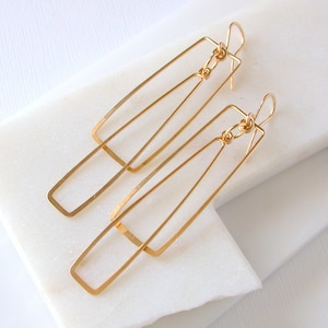 May include: Gold geometric dangle earrings with a long, rectangular shape. The earrings are made of two overlapping pieces of wire, creating a modern and minimalist design.