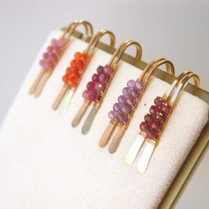 May include: Five handmade hair clips with gold-colored metal accents. Each clip features a different color of gemstone beads, including pink, orange, red, purple, and burgundy. The clips are arranged on a white surface.