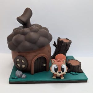 Puede incluir: Un diorama caprichoso impreso en 3D que presenta una casa en forma de bellota marrón con una pequeña puerta y una ventana redonda. Una linda ardilla de dibujos animados está de pie junto a la casa, con un tocón de árbol y rocas sobre una base verde.
