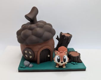 May include: A whimsical, 3D-printed diorama featuring a brown acorn-shaped house with a small door and round window. A cute cartoon squirrel stands beside the house, with a tree stump and rocks on a green base.