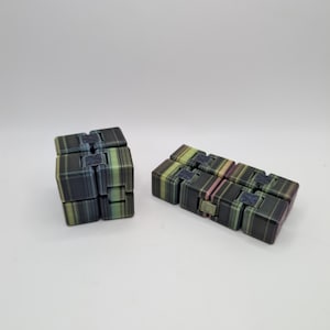 May include: Two multi-colored infinity cubes are displayed against a white background. The cubes are made of small, rectangular blocks in shades of black, green, and pink. One cube is in a cube shape, and the other is in a straight line.