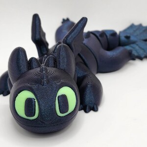 May include: A black and green 3D printed dragon figurine with glowing green eyes. The dragon is lying on its side with its wings spread out.