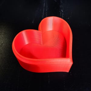 May include: A bright red heart-shaped container. The container has a smooth, curved design with a slightly textured interior. The heart shape is clearly defined, and the color is a vibrant, solid red.