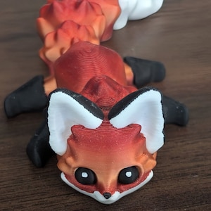 以下が含まれることがあります： 黒と白のディテールが施された、3Dプリントされた赤いキツネの置物。キツネは大きな目と長いセグメントされた尻尾を持っています。