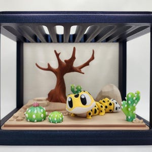Puede incluir: Una escena desértica en miniatura en un marco de madera azul oscuro. La escena incluye un gecko amarillo y negro, cactus, un árbol y una roca. El fondo es de color beige claro.