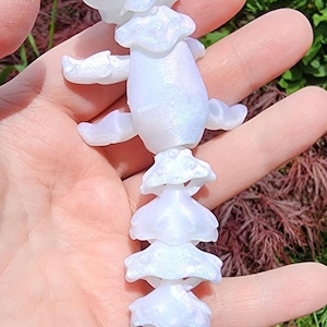 May include: A white iridescent plastic figurine of a creature with a segmented tail. The creature has a rounded head with a small, pointed snout and large, round eyes. The tail is made up of several segments, each with a slightly different shape.