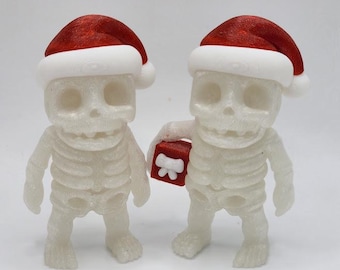 May include: Two white skeleton figurines wearing red and white Santa hats. One skeleton holds a small red gift box with a white bow. The figurines have detailed rib cages and skull faces, perfect for holiday decor.