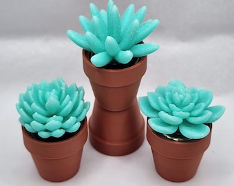 May include: Three artificial succulents in terracotta pots. The succulents are a vibrant turquoise color, with detailed petal structures. The pots vary in size and shape, creating a visually appealing arrangement. The background is a plain white.