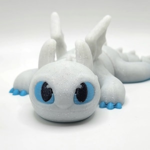 May include: A white, sparkly, 3D printed figurine of a dragon with blue eyes. The dragon is lying on its belly with its wings spread out.