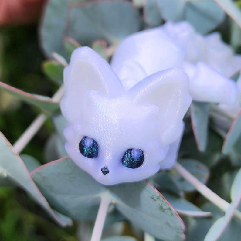 May include: A small, white, iridescent figurine of a fox with blue eyes. The figurine is sitting on a bed of green leaves.