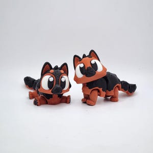 May include: Two 3D-printed toy cats in a playful pose. The cats have a black and orange color scheme with large white eyes. One cat is lying down, while the other is standing. The toys are set against a plain white background.