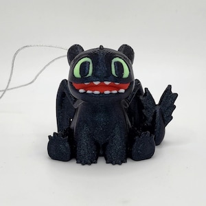May include: Black and green 3D printed ornament of a cartoon dragon with a toothy grin. The dragon has a string loop for hanging.
