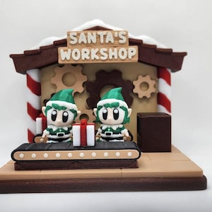 May include: A festive Christmas decoration featuring Santa's Workshop. Two elf figures in green and white outfits stand near a conveyor belt with gifts. The workshop has a brown roof with the text "SANTA'S WORKSHOP".