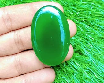 May include: A polished, oval-shaped, jade-green gemstone. The stone is held in a hand, with a bright green, grassy background. The gemstone has a smooth, reflective surface, and is a vibrant shade of green.