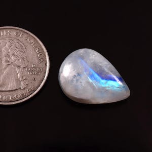 Può includere: Una pietra di luna lucidata a forma di pera, con una base bianco latte e un bagliore blu iridescente. Una moneta da un quarto di dollaro USA è accanto alla gemma per il confronto delle dimensioni. La pietra è su uno sfondo nero.