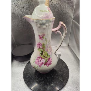 Puede incluir: Una jarra de porcelana blanca con tapa, con rosas rosas y hojas verdes. La jarra tiene un asa curva y un pico, con detalles dorados. El diseño está pintado a mano y la jarra es probablemente vintage.