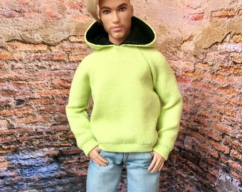 Könnte beinhalten: Puppe mit einem limettengrünen Kapuzenpullover mit schwarzem Kapuzenfutter, hellblauen Jeans und weißen Turnschuhen. Die Puppe hat blondes Haar und steht vor einer Backsteinmauer.