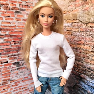 以下が含まれることがあります： 白い長袖シャツと青いジーンズを着た人形。人形は長い金髪で、フープイヤリングをしています。背景はレンガの壁です。
