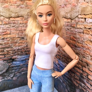 Könnte beinhalten: Eine Puppe mit blondem Haar, die ein weißes Tanktop und hellblaue Leggings trägt. Die Puppe hat blaue Augen und trägt silberne Ohrringe. Der Hintergrund ist eine Backsteinmauer.