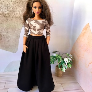 May include: A doll dressed in a two-piece ensemble. The top is a cropped, off-the-shoulder design with a white and brown cow print. The doll is also wearing a long, black pleated skirt. The doll has long brown hair and is wearing hoop earrings.