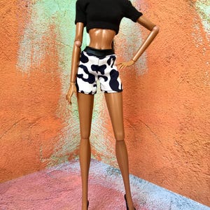 May include: A fashion doll with a dark complexion wearing a black short-sleeved crop top and white shorts featuring a black cow print pattern. The doll has long dark hair, silver hoop earrings, and is wearing black high-heeled shoes on its feet.