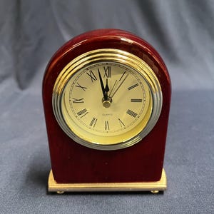 Peut inclure: Une petite horloge à quartz de couleur marron foncé avec une base dorée. Le cadran est crème avec des chiffres romains et des aiguilles noirs. L'horloge mesure environ 10 cm de haut.