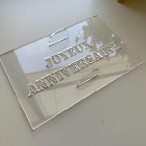 May include: A clear acrylic plaque with the words "JOYEUX ANNIVERSAIRE" in raised lettering. The plaque has a rectangular shape with rounded corners and two oval-shaped decorative elements.