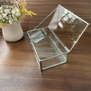 May include: A clear glass box with a silver metal frame and a hinged lid. The lid is held closed with a silver chain.