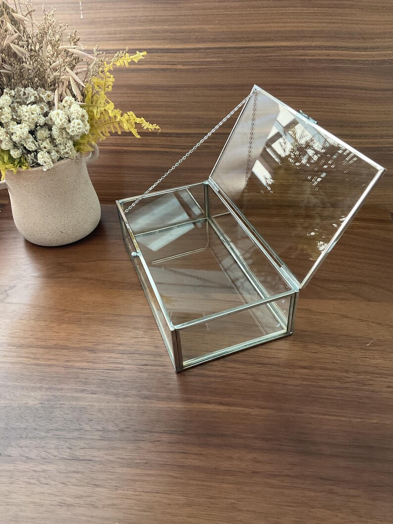 May include: A clear glass box with a silver metal frame and a hinged lid. The lid is held closed with a silver chain.