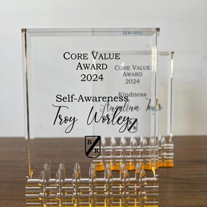 May include: Clear acrylic award with a gold base. The text "Core Value Award 2024" and "Self-Awareness" are printed on the front. The name "Troy Worley" is also displayed. The award has a decorative, textured base.