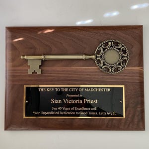 May include: A wooden plaque with a gold key and a black plaque with gold lettering. The plaque reads "The Key to the City of Madchester. Presented to Sian Victoria Priest. For 40 Years of Excellence and Your Unparalleled Dedication to Good Times. Let's Ave It."