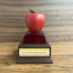 May include: A red apple trophy with a wooden base. The base has a gold plaque with the text "Your Custom Engraving Here".