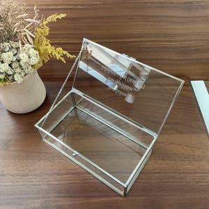 May include: A clear glass box with a silver metal frame and a hinged lid. The box has a chain attached to the lid. The box is engraved with the text "Congratulations on your Graduation 2022 from Jackson High School".