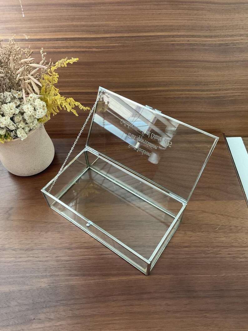 May include: A clear glass box with a silver metal frame and a hinged lid. The box has a chain attached to the lid. The box is engraved with the text "Congratulations on your Graduation 2022 from Jackson High School".