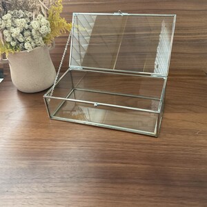 May include: A clear glass display box with a silver metal frame and a hinged lid. The lid is attached to the box with a silver chain.