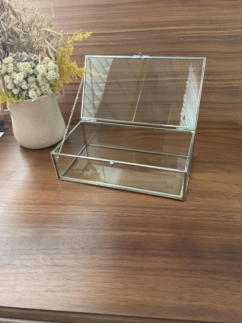 May include: A clear glass display box with a silver metal frame and a hinged lid. The lid is attached to the box with a silver chain.