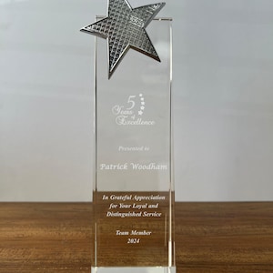 May include: A clear rectangular glass award trophy topped with a silver-toned star featuring a textured grid pattern. The glass is etched with the text 5 Years of Excellence, Presented to Patrick Woodham, In Grateful Appreciation for Your Loyal and Distinguished Service, Team Member 2024.