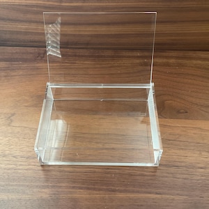 May include: A clear acrylic display box with a hinged lid. The box is rectangular and sits on a wooden surface. The lid is upright, showing the interior of the box. The display box is designed to showcase small items.