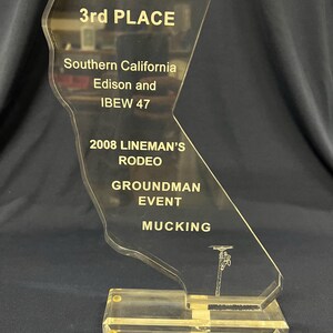 May include: A clear acrylic trophy in the shape of California, with engraved text. The trophy reads "3rd PLACE", "2008 LINEMAN'S RODEO", and "GROUNDMAN EVENT MUCKING". The base is rectangular and clear.