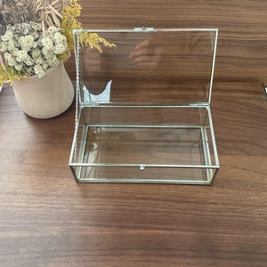 May include: A clear glass box with a silver metal frame and a hinged lid. The box is open and the lid is propped up.