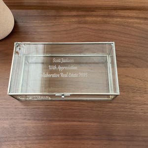 May include: A clear glass box with a silver trim and a hinged lid. The box is engraved with the text "Scott Jackson, With Appreciation, Collaborative Real Estate 2025".