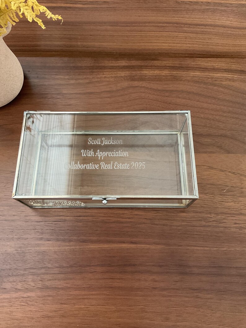 May include: A clear glass box with a silver trim and a hinged lid. The box is engraved with the text "Scott Jackson, With Appreciation, Collaborative Real Estate 2025".