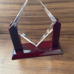 May include: A clear crystal award with a diamond-shaped design, set on a rectangular, dark red wooden base. The award features a polished finish and is supported by two matching red wooden pillars.