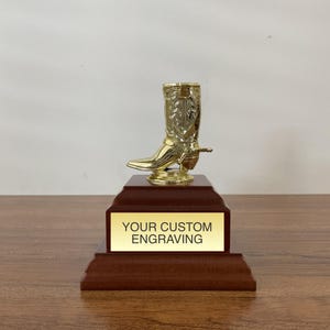 May include: A gold-colored cowboy boot trophy topper mounted on a tiered dark brown wooden base. A rectangular gold plaque is attached to the front of the base with the black text "YOUR CUSTOM ENGRAVING" printed clearly in the center of the plate.